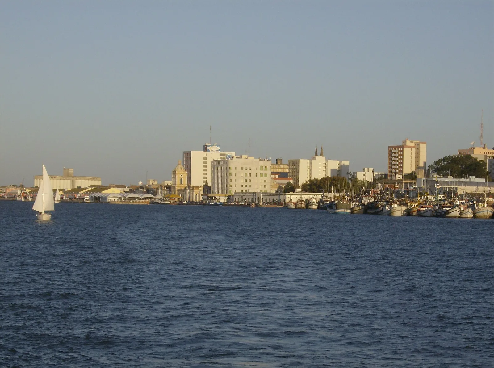 Rio Grande (RS) vista da água: o cais com os barcos de pesca, os prédios do centro e um veleiro na Lagoa dos Patos
